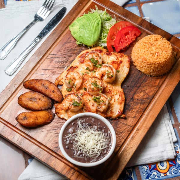 Mexican chicken and shrimp dish with rice, beans, plantains, avocado, and tomatoes on a wooden board.