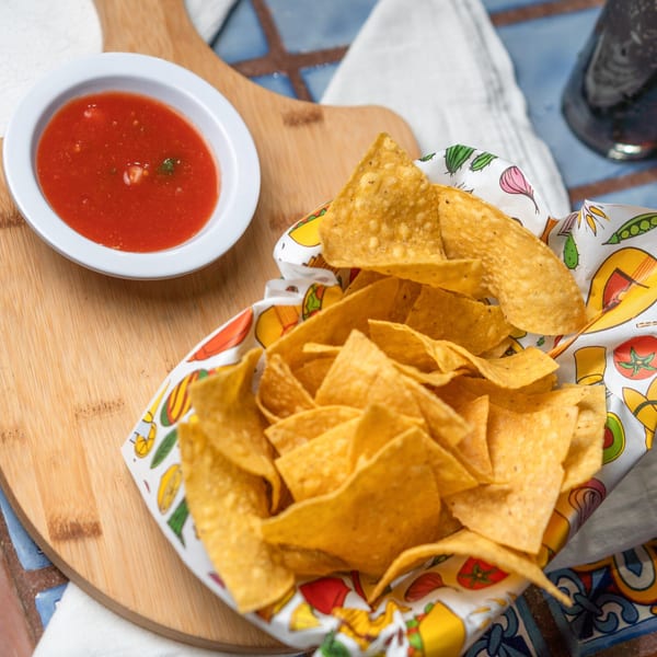 A basket of tortilla chips and a white bowl of red salsa on a wooden board.