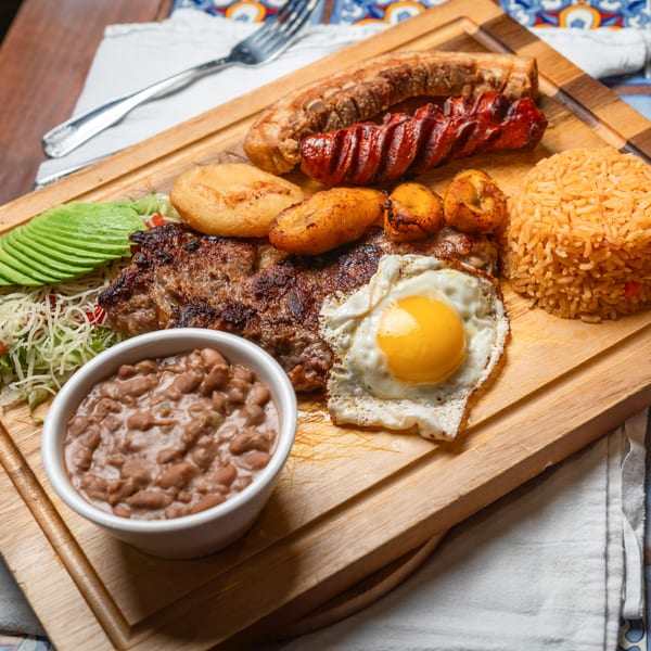 A Mexican platter with steak, chorizo, fried egg, rice, beans, avocado, and plantains on a wooden board.