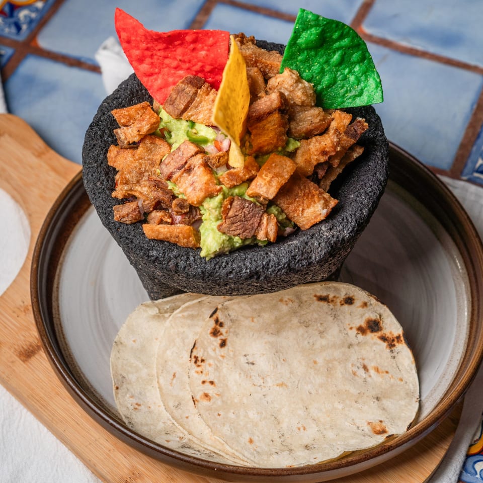Guacamole with chicharrones and colorful tortilla chips in a molcajete, served with tortillas.