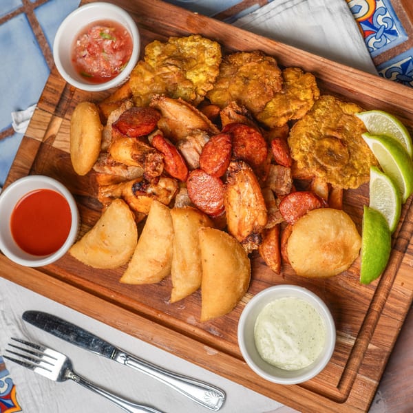 A wooden board with fried plantains, empanadas, sausage, chicken, and three dipping sauces.